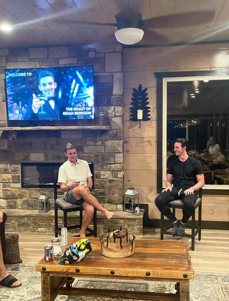 A wall mounted TV reading 'The Roast of Noah Berkson' above a stone fireplace, with two guests seated on stools at a cabin gathering