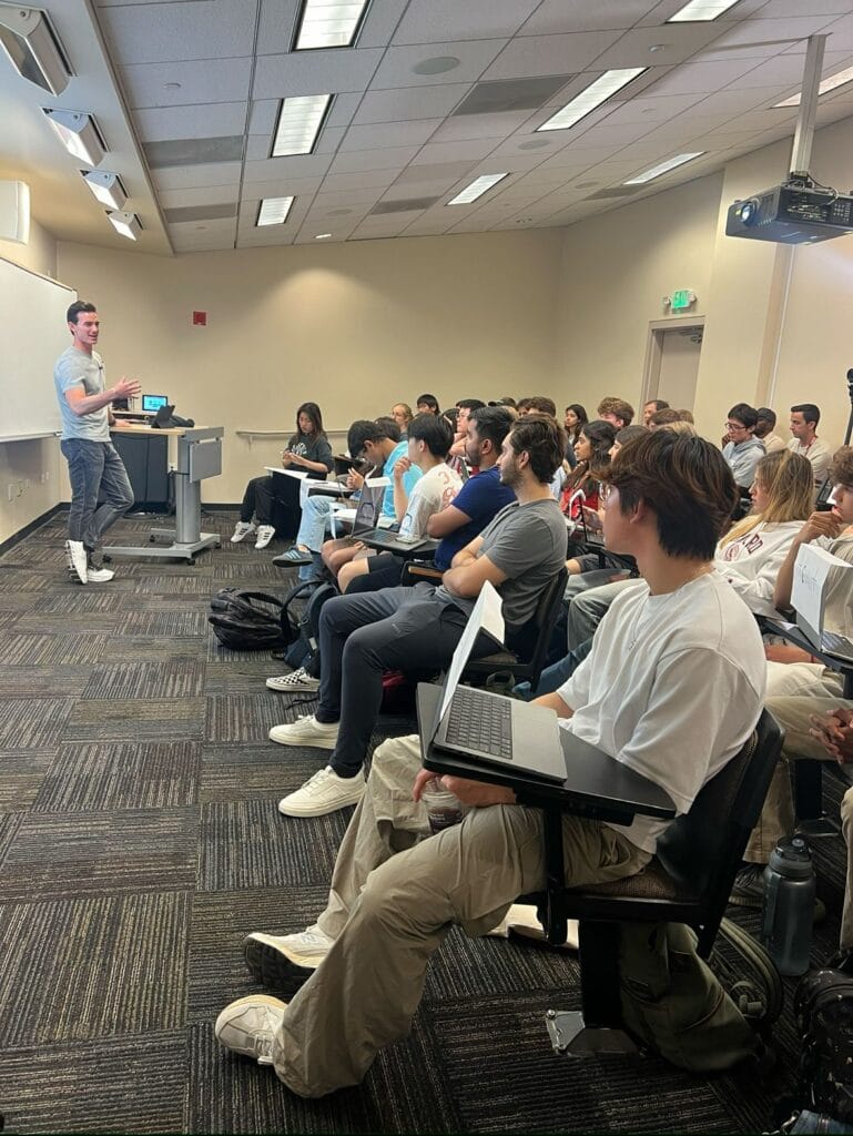 Noah Berkson speaking to a room of seated students with laptops in a university classroom